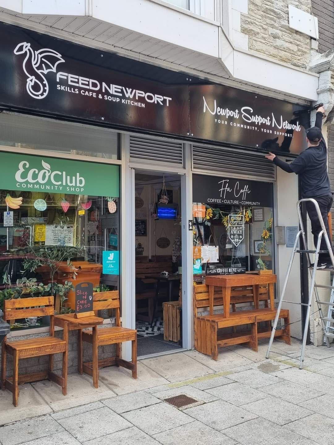 Storefront of Feed Newport Skills Cafe & Soup Kitchen and Newport Support Network with wooden benches outside.