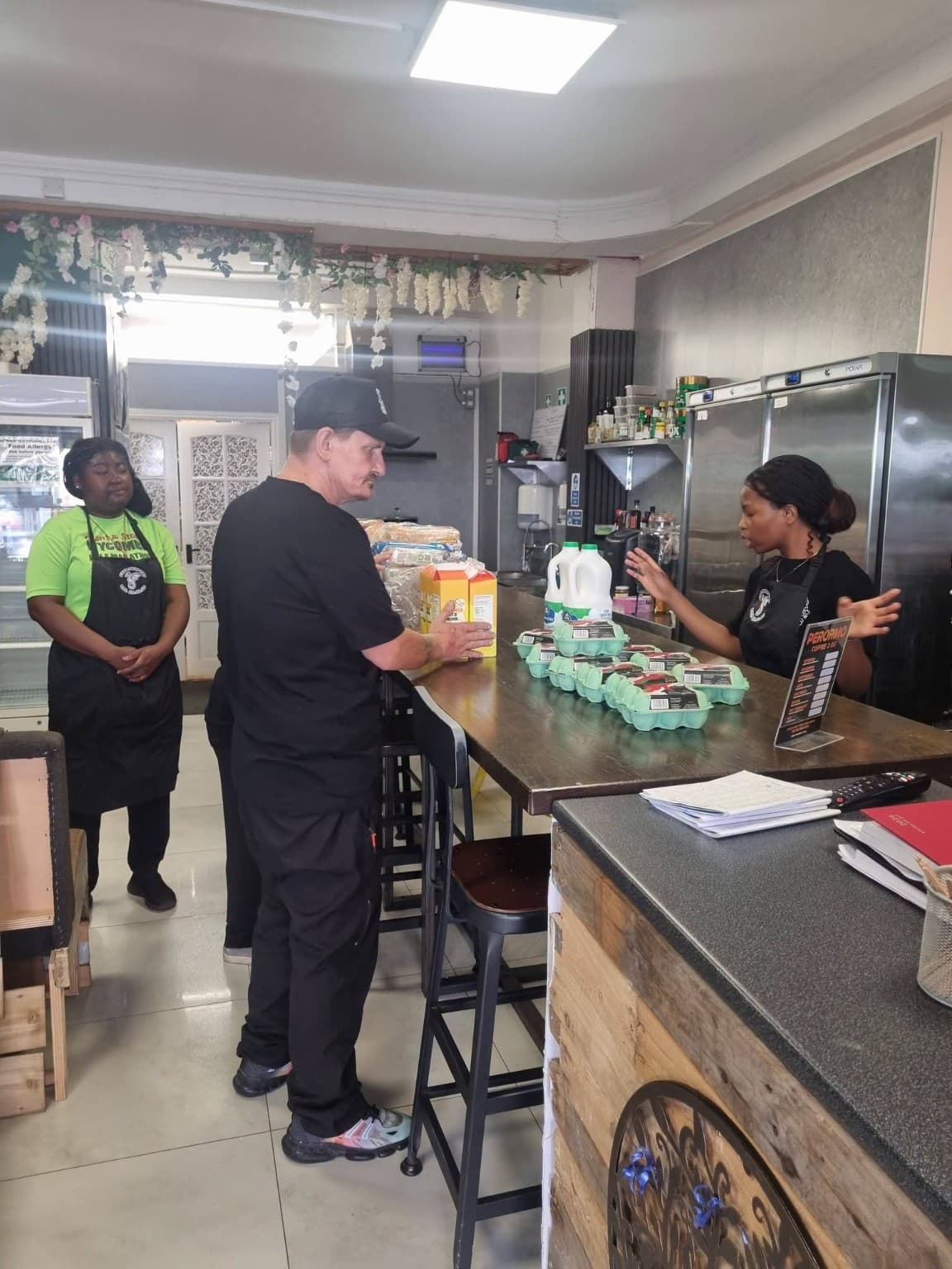 A man places groceries on a cafe counter while two staff members in aprons watch.