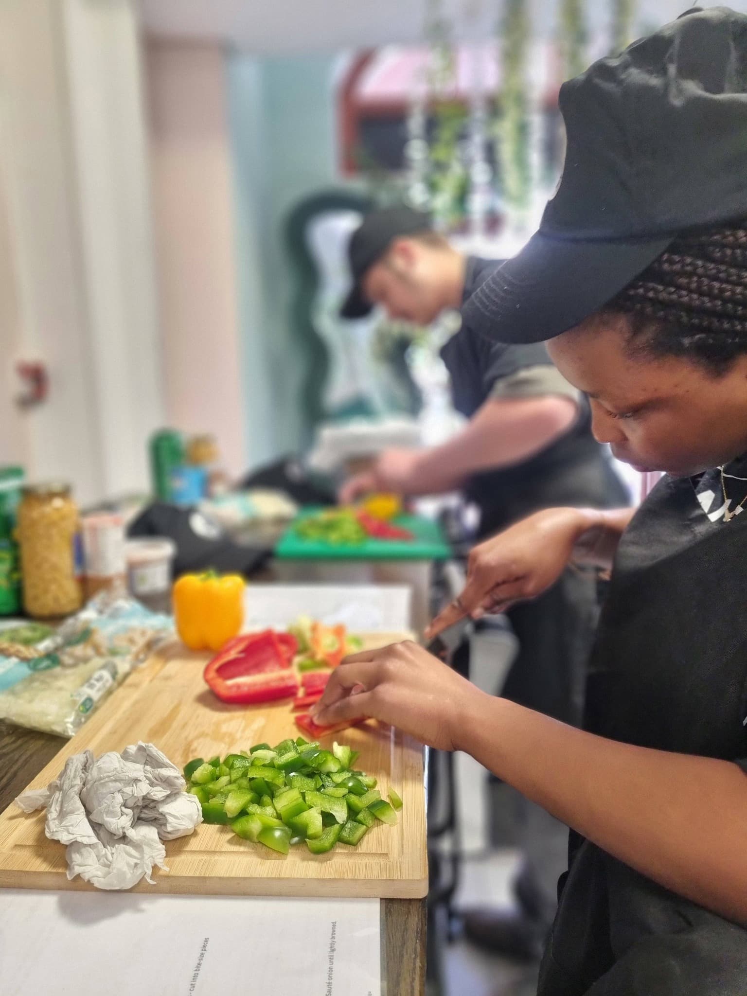 Two people in black uniforms chop colorful bell peppers on cutting boards in a kitchen.