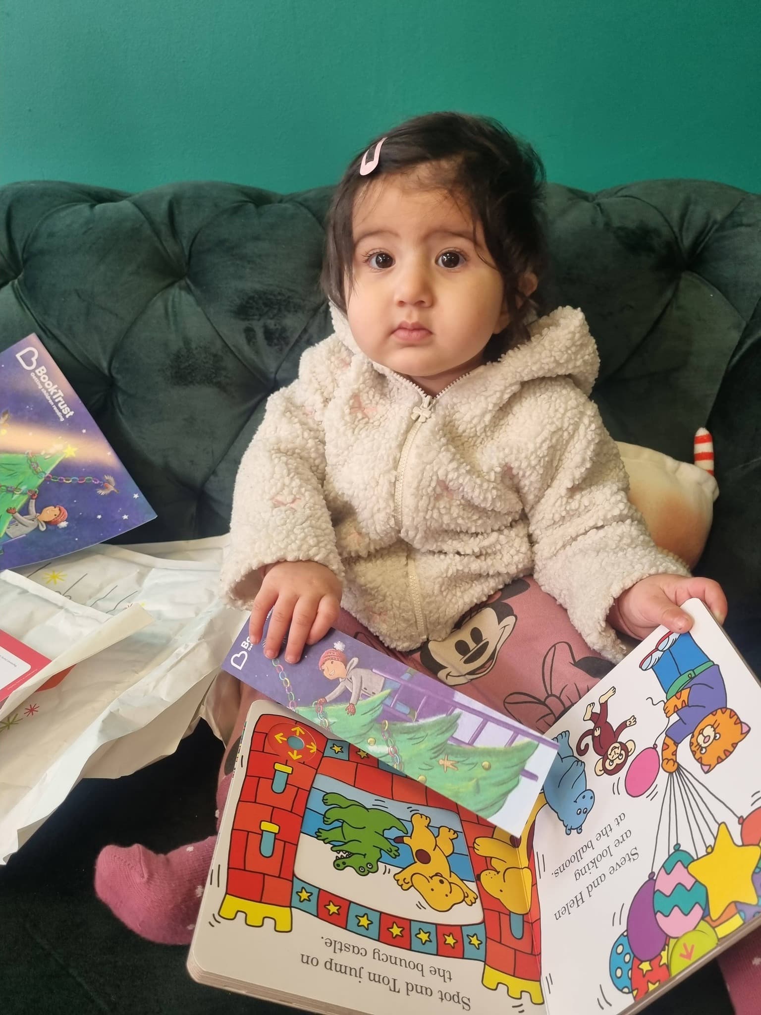Toddler in a white sherpa hoodie sits on a green sofa holding an open book.