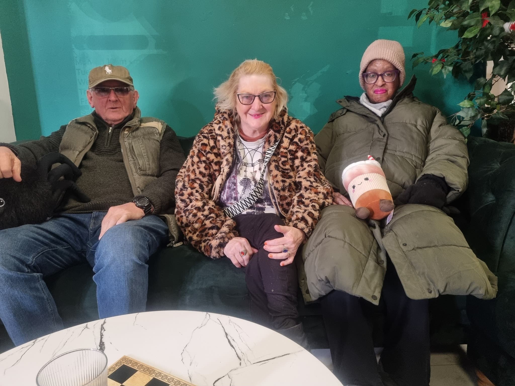 Three elderly people sit together on a green sofa, wearing warm winter coats and glasses.