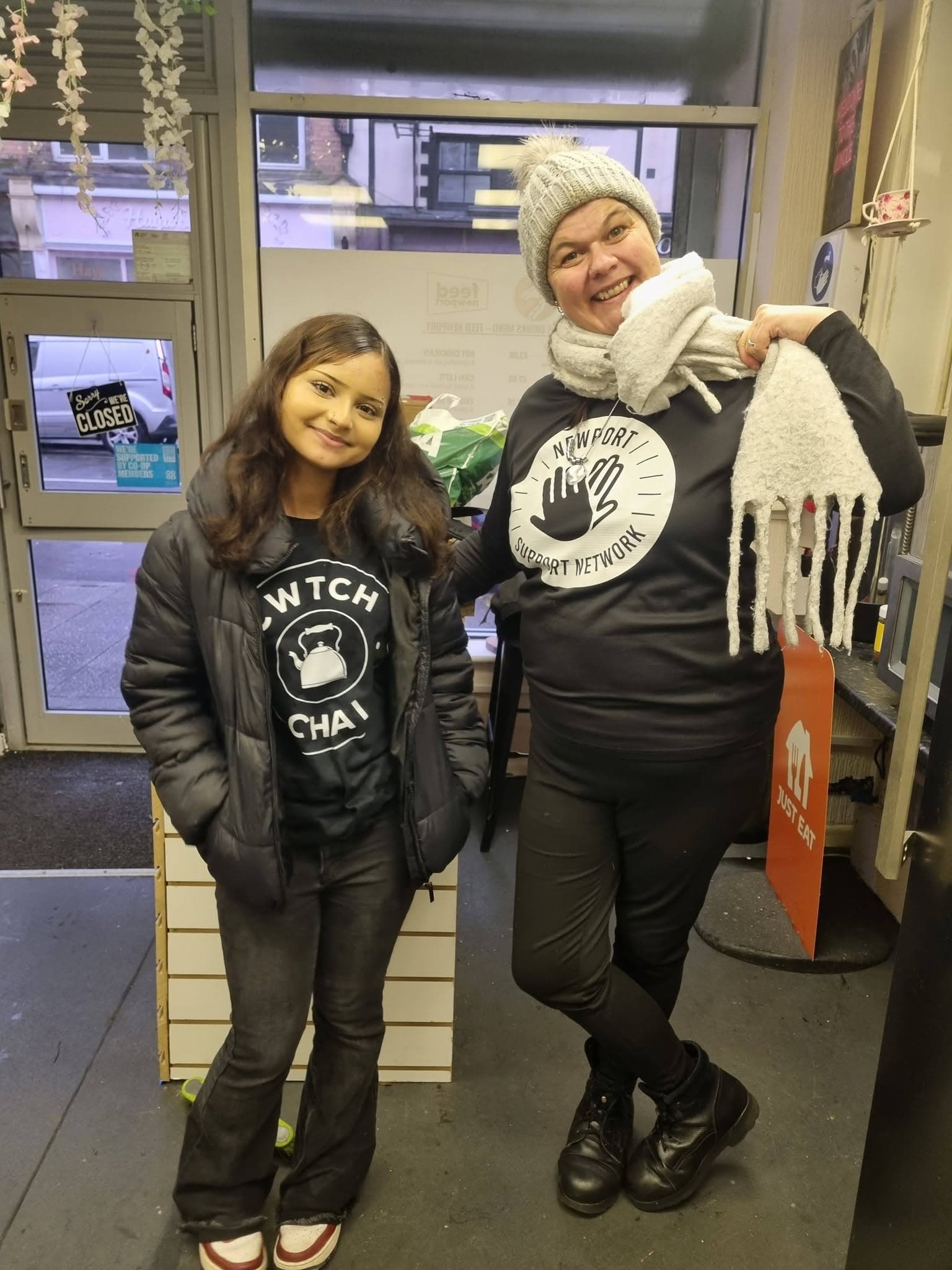 Two smiling women pose indoors wearing 'Witch Chai' and 'Newport Support Network' branded apparel.