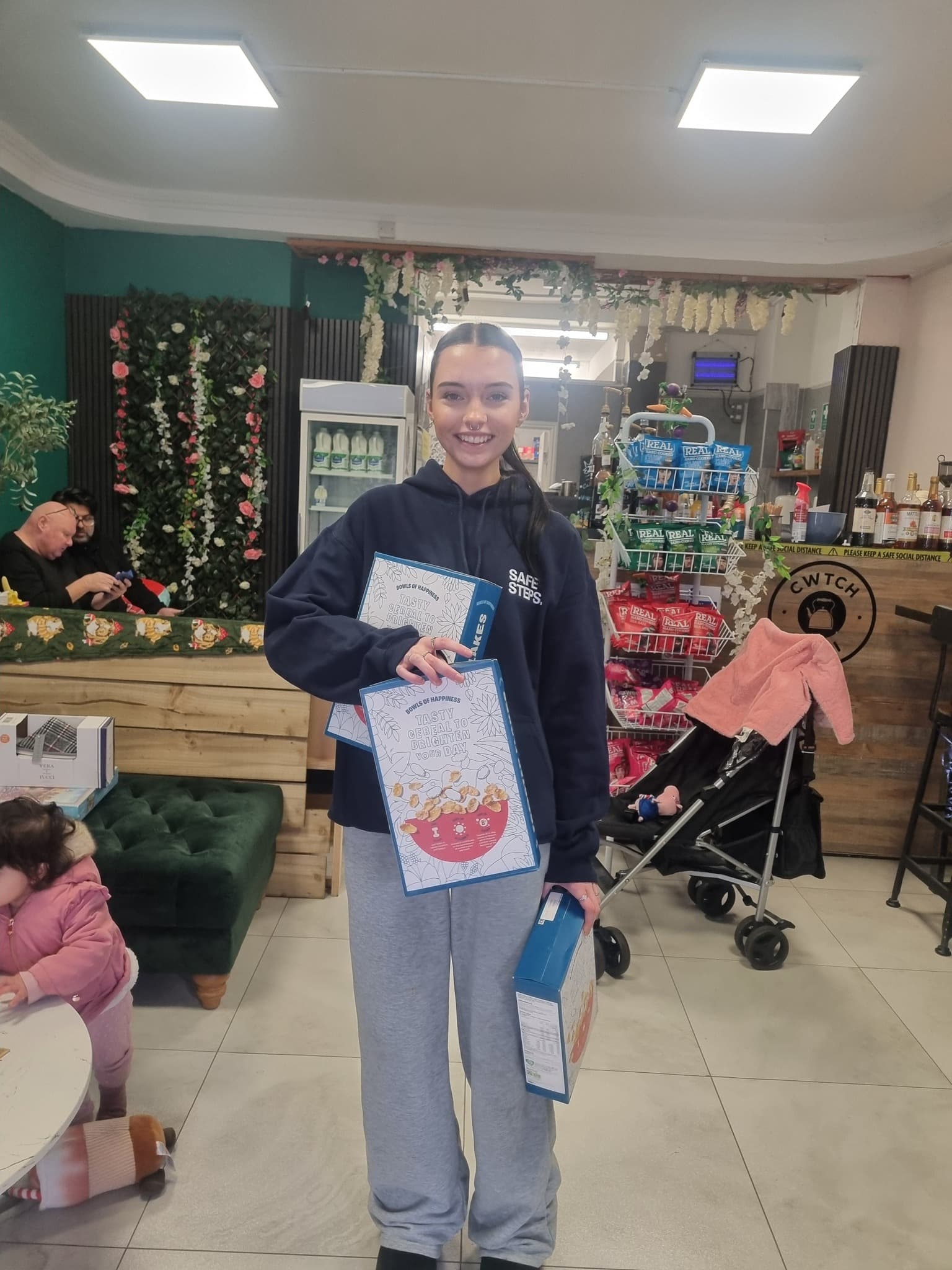 Smiling woman in a hoodie holding three cereal boxes inside a cafe with floral decorations.