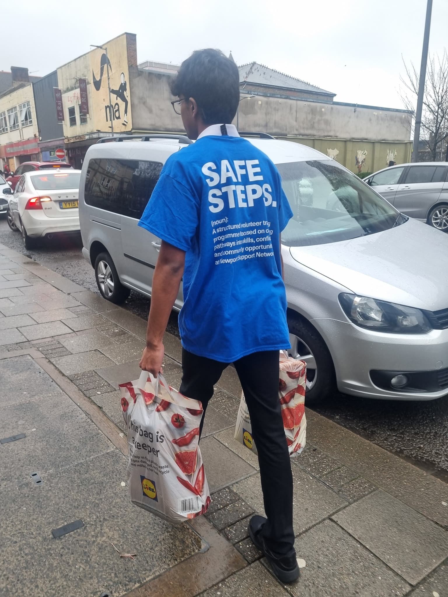 Young man in a blue Safe Steps shirt carries grocery bags on a city sidewalk.