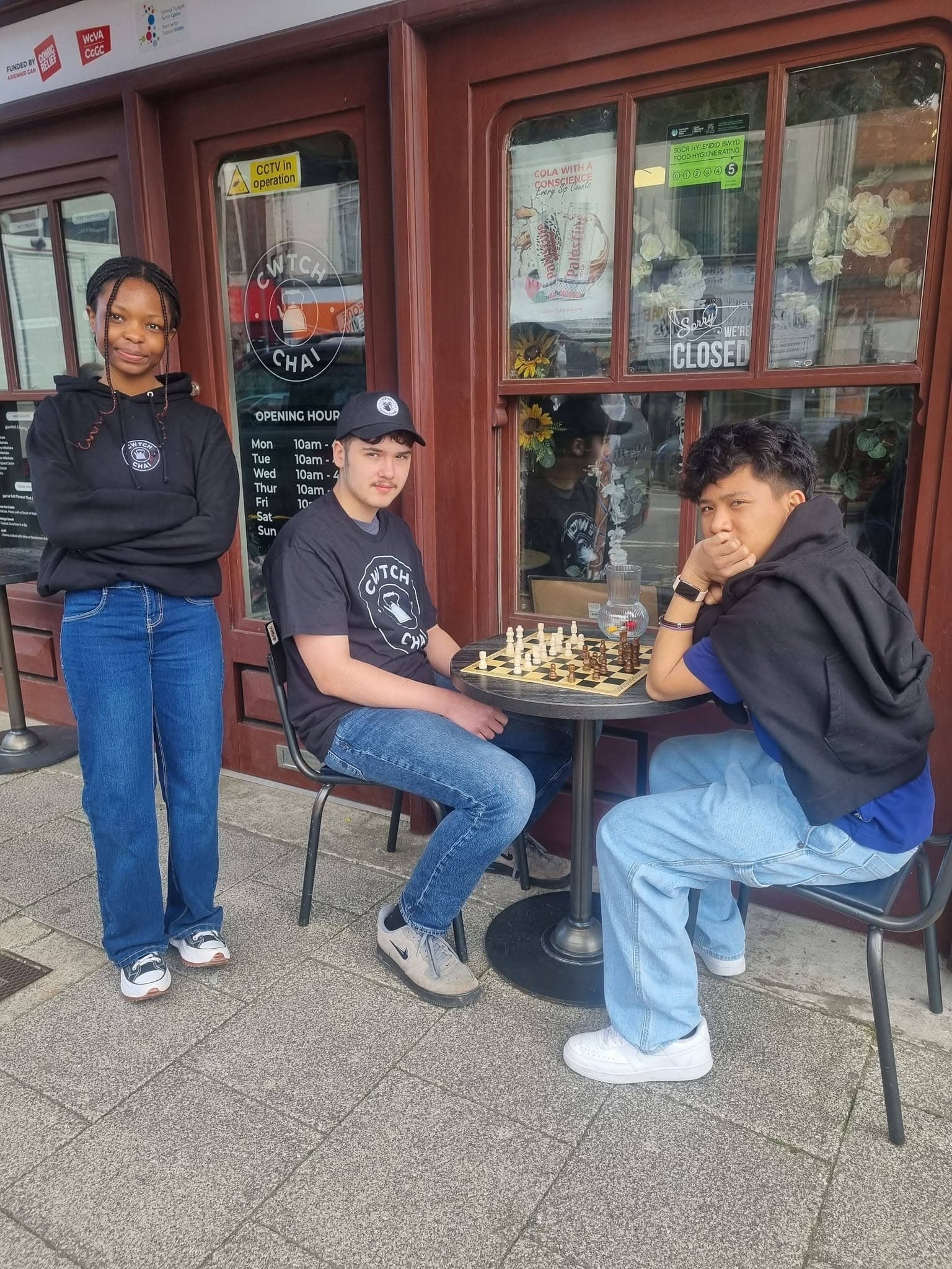 Three young people outside Cwtch Chai cafe, two seated at a table playing chess.