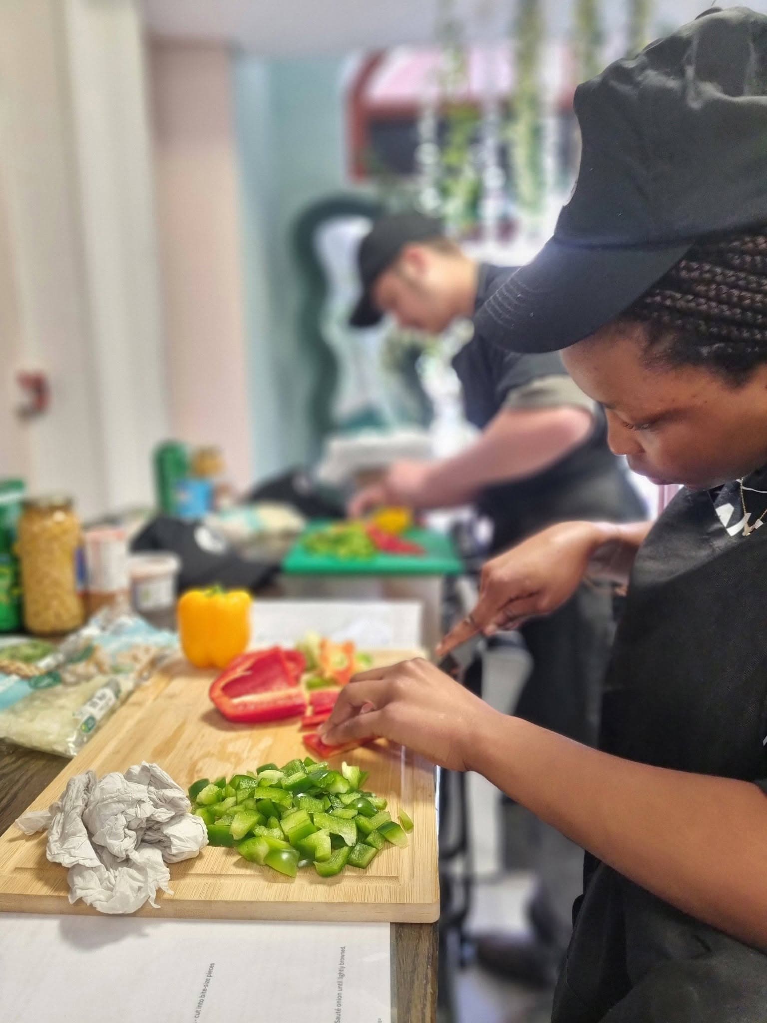 Two cooks in black caps chop bell peppers on cutting boards in a kitchen.