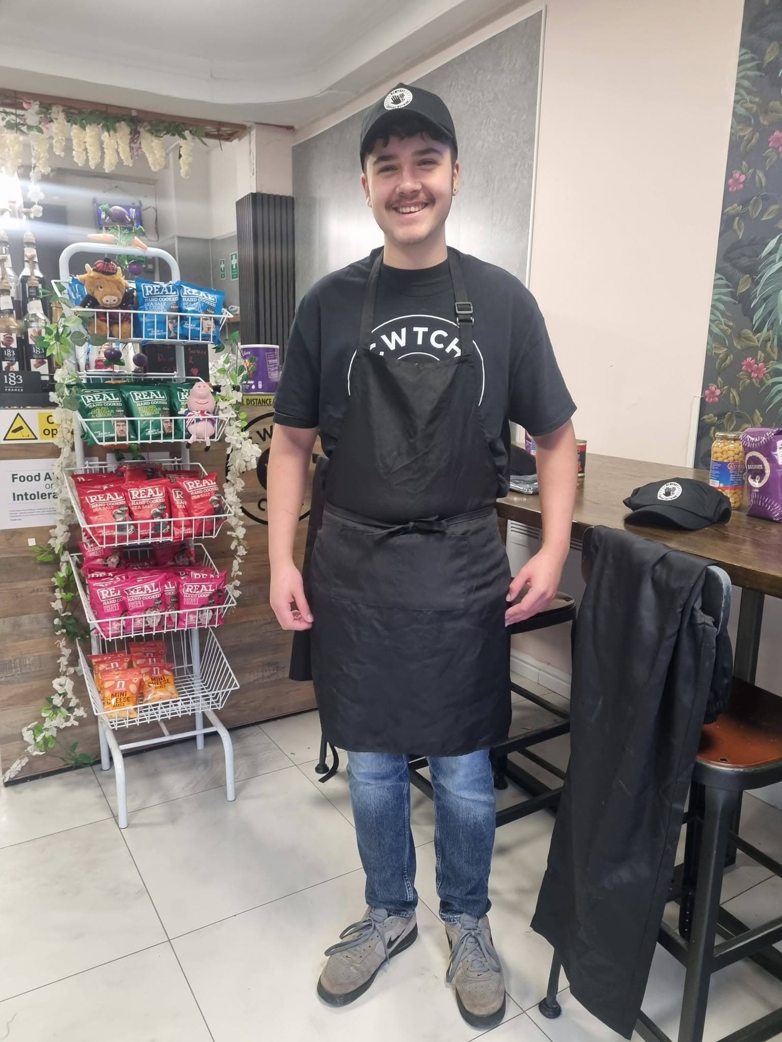 Smiling young man in a black apron and cap standing in a cafe with snacks.