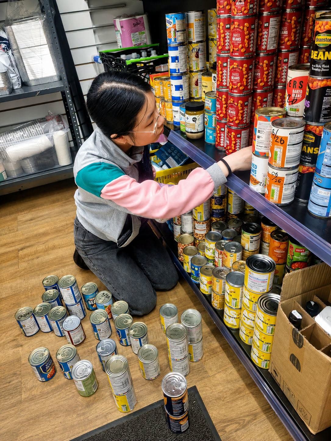 Woman in a colorful jacket kneels on the floor, organizing canned goods on pantry shelves.